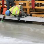 Image is of two construction workers in neon vests sitting on a platform above freshly poured concrete, which they are working on treating.