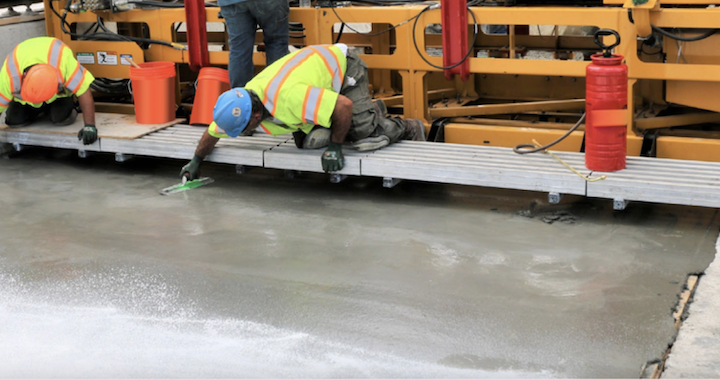 Image is of two construction workers in neon vests sitting on a platform above freshly poured concrete, which they are working on treating.