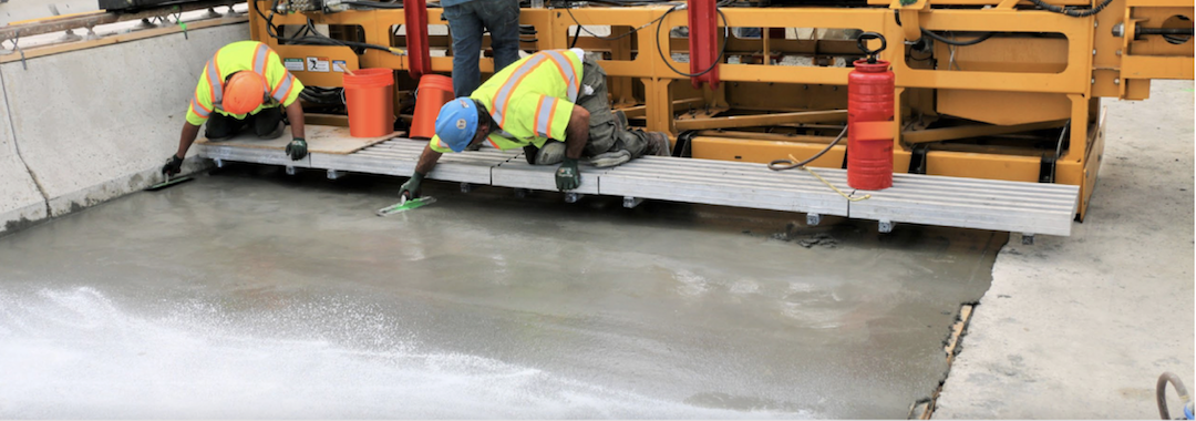 Image is of two construction workers in neon vests sitting on a platform above freshly poured concrete, which they are working on treating.