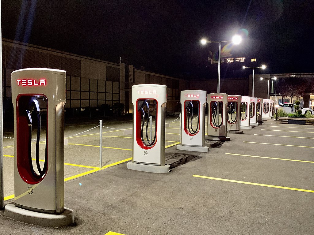 Image of a row of Tesla Superchargers in a parking lot. The chargers are rectangular with plugs resembling gas pumps inside the hollow rectangle.