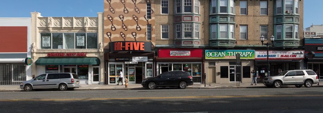 Image of a street iwth four lanes for traffic, three parked cars, and a series of shops, such as center city deli, hi five, Ocean Therapy, and casino city barber and salon