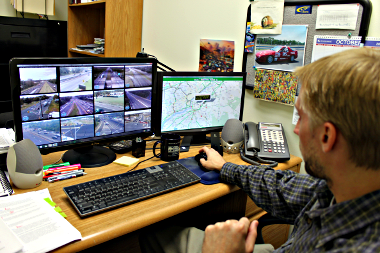 Image of a man looking at a two computer screens: the one on the left is of highway traffic cameras, and the right shows a map of the region's roads and their status, green for no traffic, red for bad traffic.