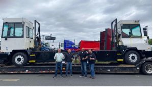 Four men stand smiling in front of a trailer with two small white truck-like vehicles on them, the electric yard tractors that were just delivered to this facility.