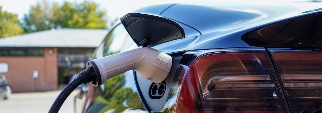 Image of a black car with a white electric charger plugged in to the rear left of it, next to the tail light.