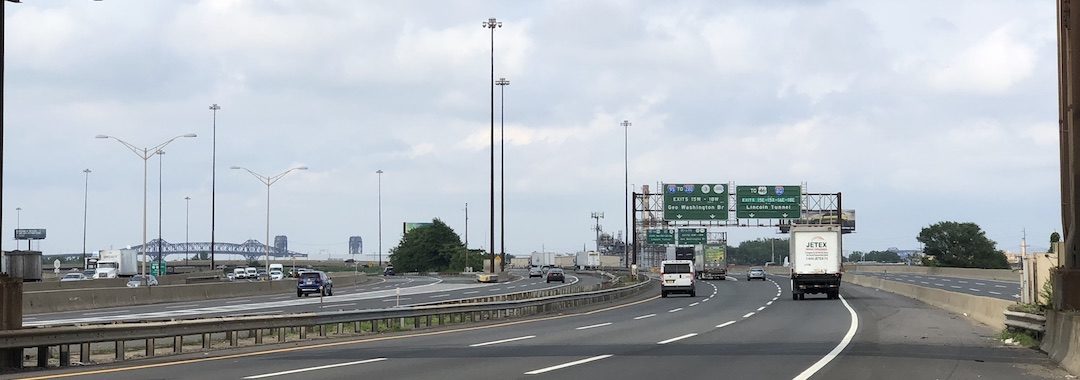 Image of a highway with two cars driving on it, in the distance the erector-set outline of the Pulaski Skyway can be seen.