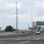 Image of a highway with two cars driving on it, in the distance the erector-set outline of the Pulaski Skyway can be seen.