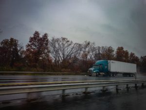 image of a blue eighteen wheeler with a long white trailer driving on a highway with trees losing their leaves on an overcast day.