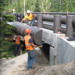 From left to right: a woman in a hardhat working with a crowbar, then two men helping place a bridge component, finally three men on a paving device