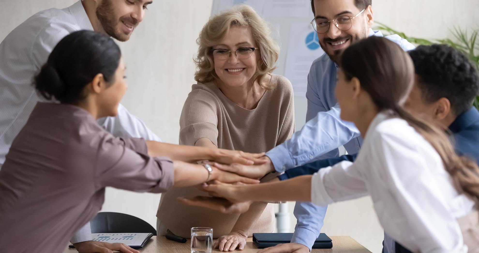 Diverse workers with mature mentor, putting hands together, showing support and unity after successful presentation at company meeting. Happy female teacher with colleagues in team building activity.