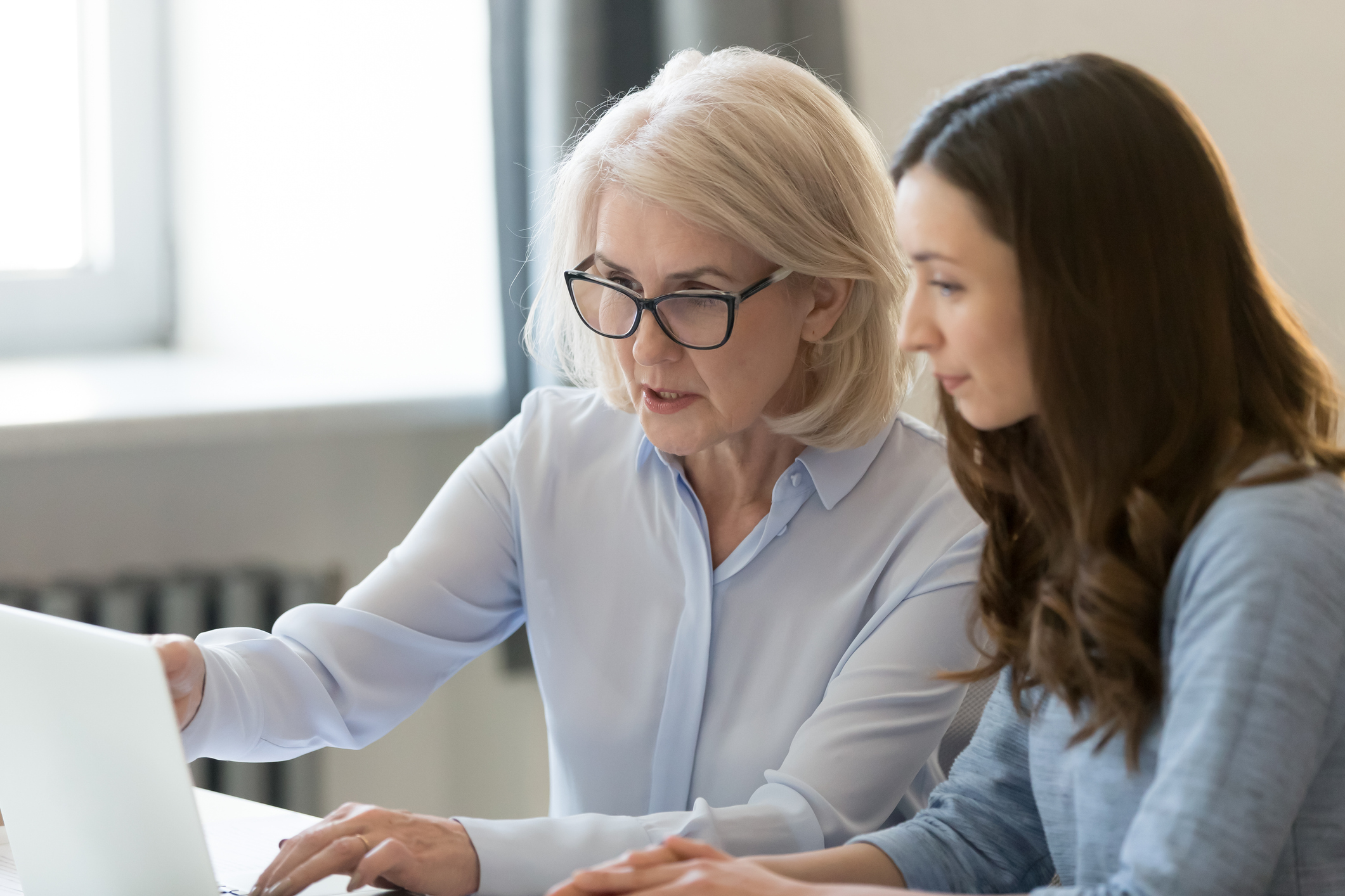 Serious old female mentor teacher coach teaching intern or student computer work pointing at laptop, mature executive manager explaining online project to young employee learning new skills in office